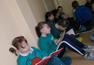 Kilkoro uczniów siedzi na korytarzu przy sali lekcyjnej i czyta książki.