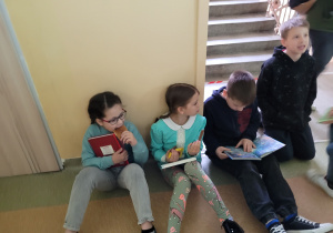 Troje uczniów czyta książki na korytarzu szkolonym przy schodach.