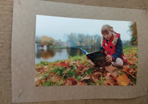 fotografia przedstawiająca dziewczynkę, która siedzi na trawie w liściach i czyta książkę